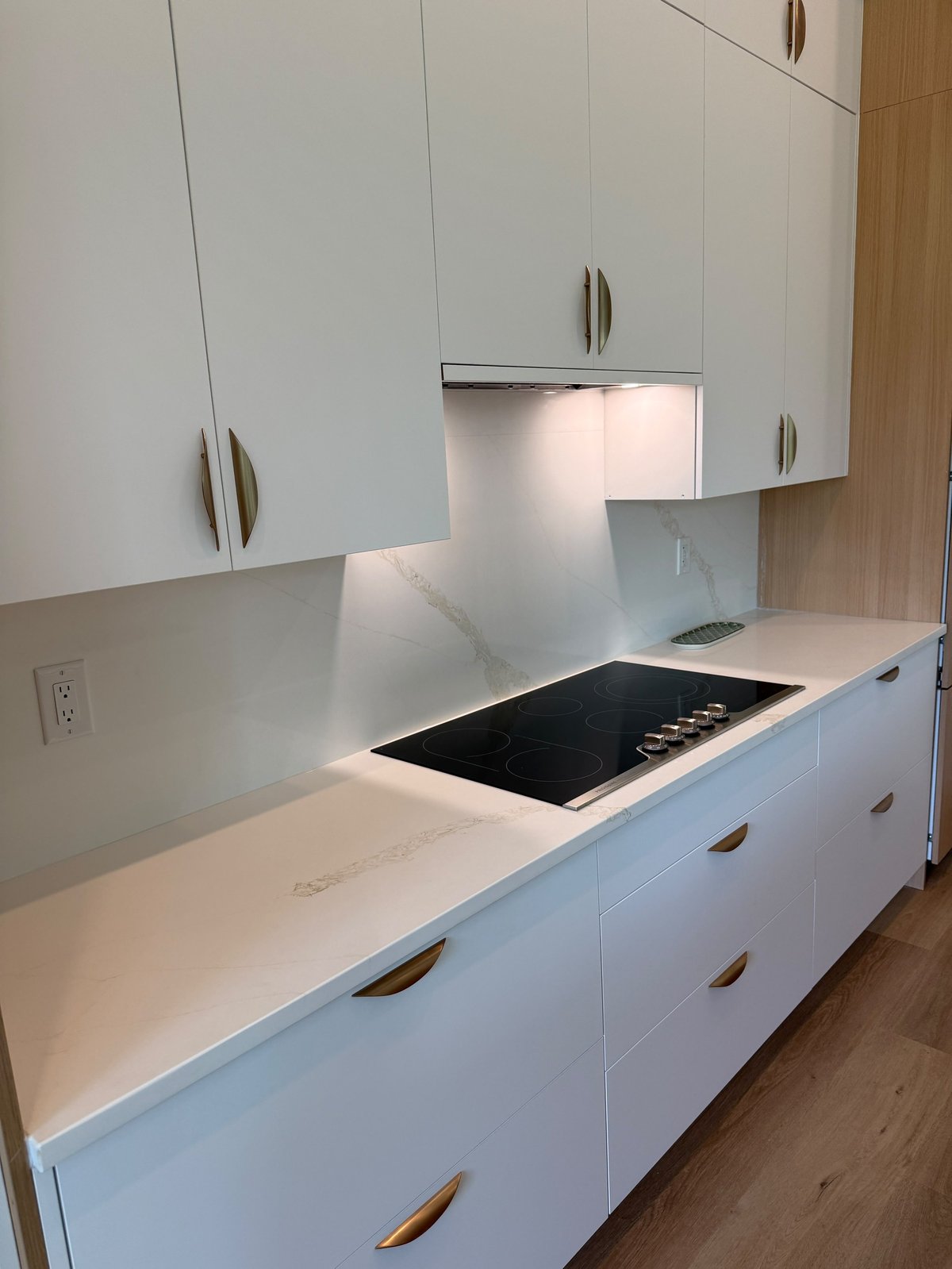 Modern white kitchen with black induction cooktop, white countertops, cabinets with bronze handles, and backsplash