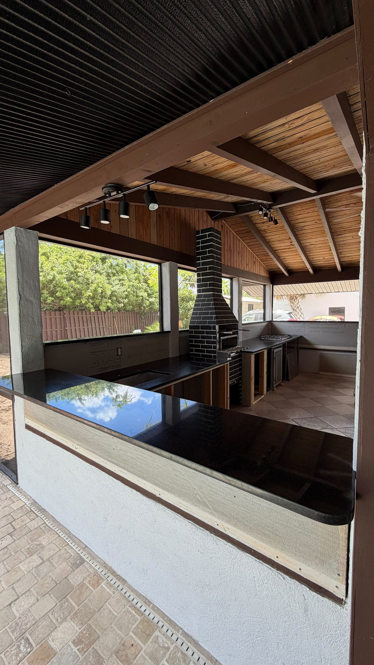 Modern outdoor pavilion with black granite countertop, brick fireplace, exposed wood beams, and black corrugated roof